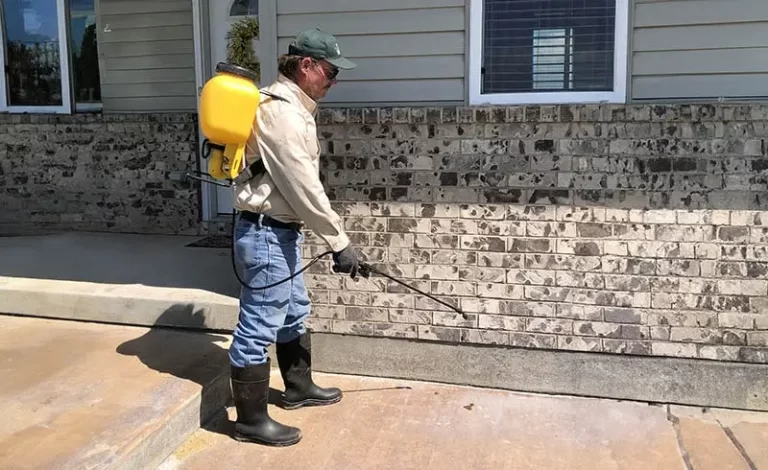 A technician with a backpack sprayer, high rubber boots, gloves, long sleeves and other appropriate PPE applies a bug spray to the outside of a house.